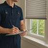 A professional installer in navy overalls writing on a clipboard near a window with modern blinds in a bright, sunlit room